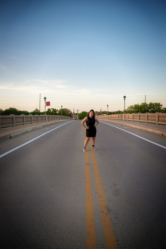 Bold urban fine art portrait of a woman in a black dress on a bridge at dusk.