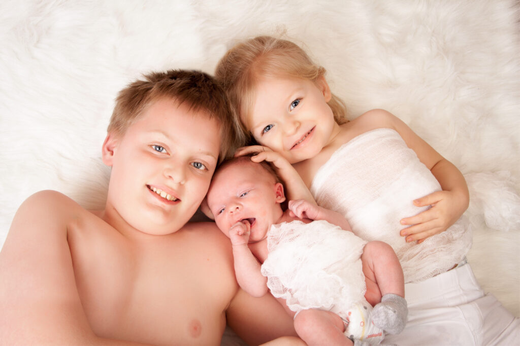 Tender fine art portrait of two older siblings cradling their newborn baby sibling on a soft white fur blanket.