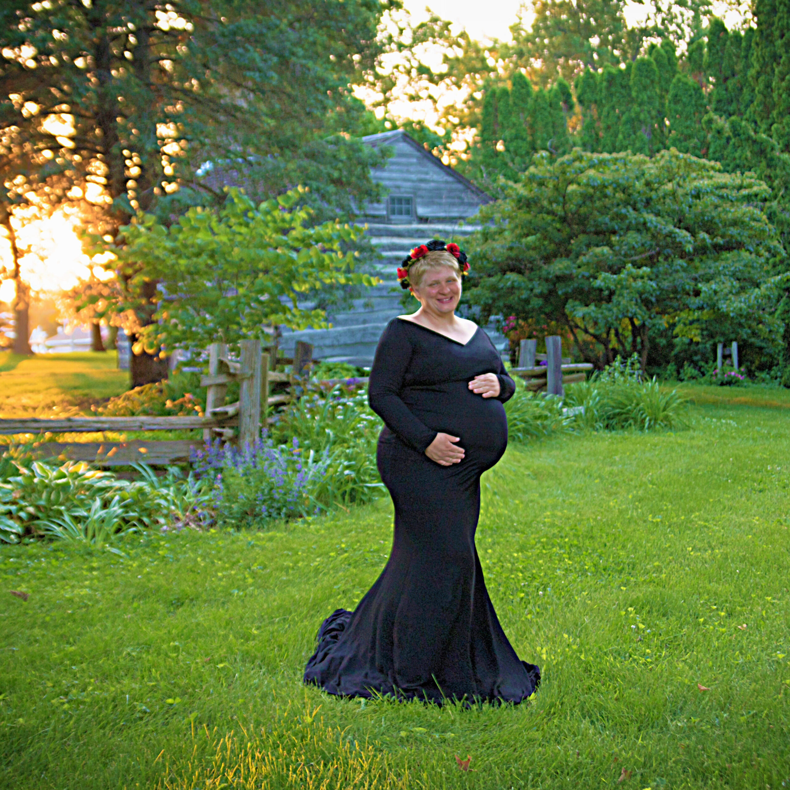 Radiant luxury maternity portrait of a woman in a black gown and floral crown at sunset in a garden setting.